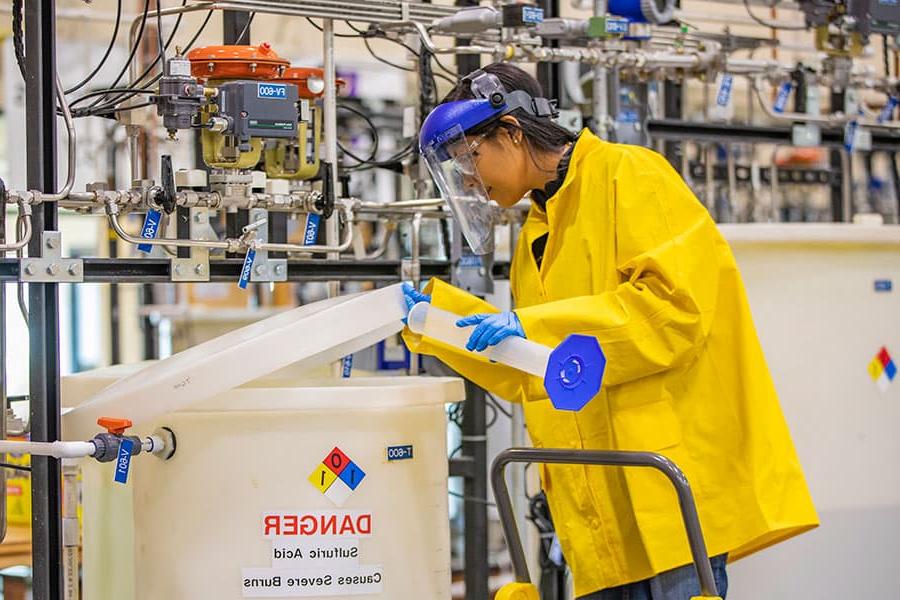 A person in a yellow lab jacket pours a beaker of solution into a container of sulfuric acid